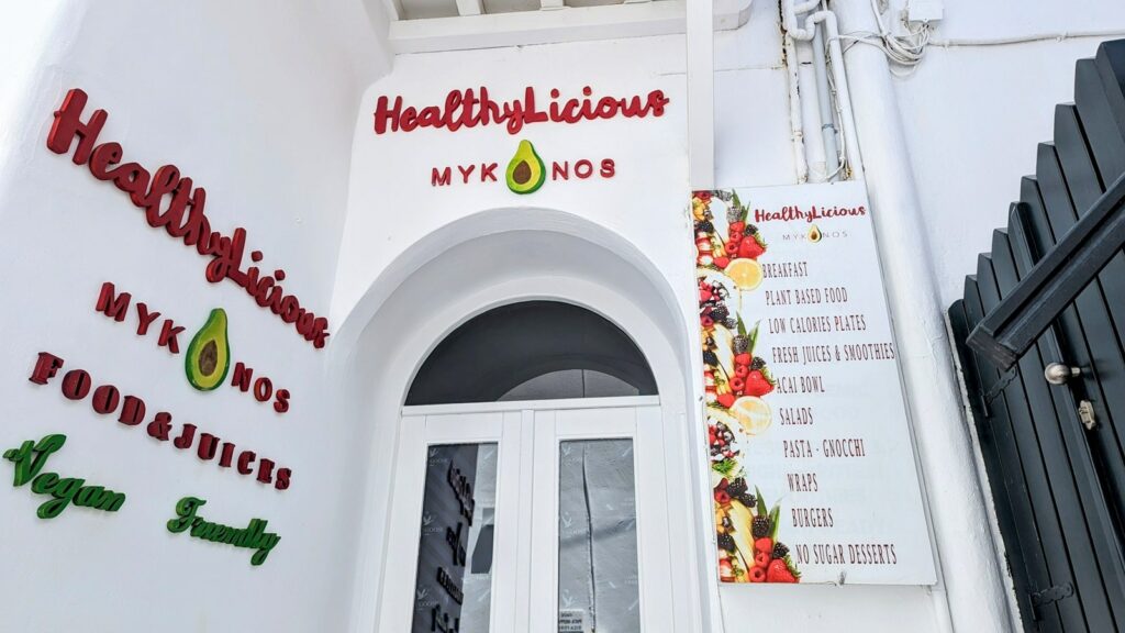the bright white outside of the vegan friendly cafe healthlicious with colorful writing listing all of their menu options and features in mykonos