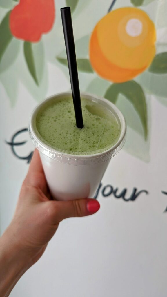 a green vegan smoothie in a white cup in front of a colorful fruit mural at juicy girl in naxos