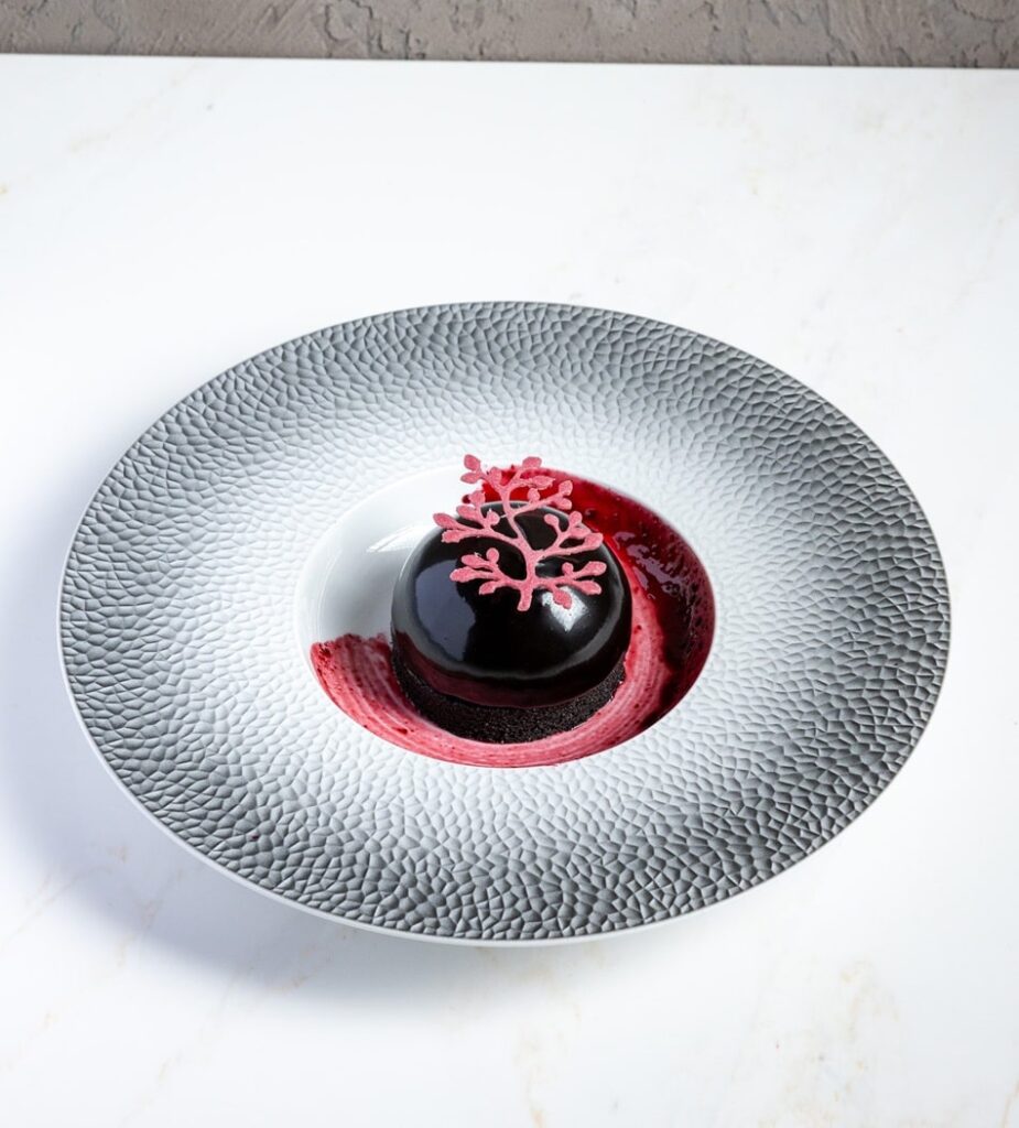 small round dark vegan cake artfully displayed in the middle of a large white dish and topped with a lacey sugar piece 