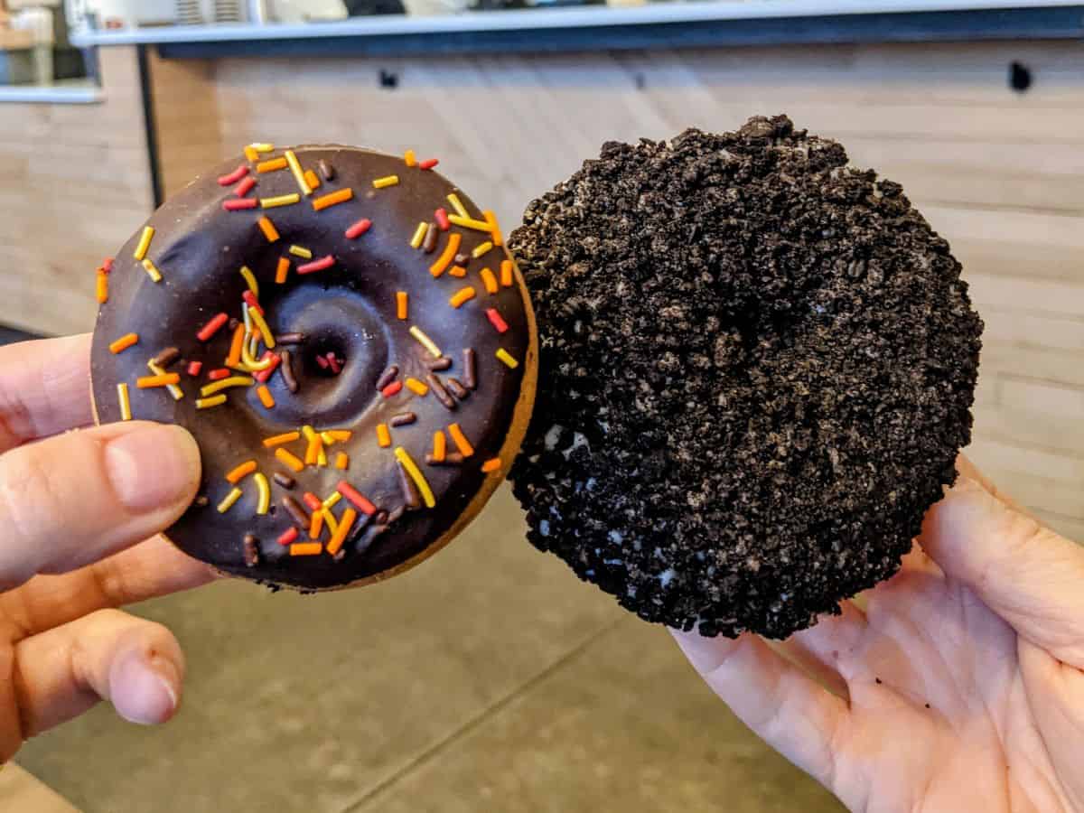 two vegan donuts covered in chocolate, cookies, and sprinkles held next to each other at a vegan bakery