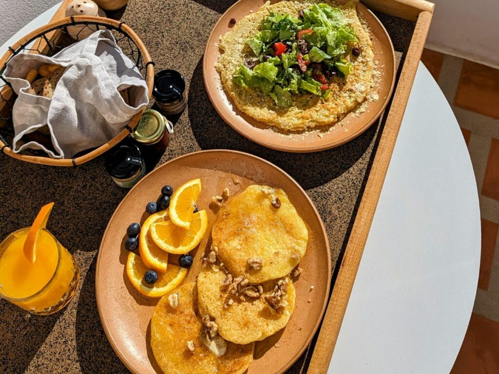 a round plate with mini vegan pancakes next to a vegan chickpea omelet on a tray with fresh juice and a bread basket in santorini