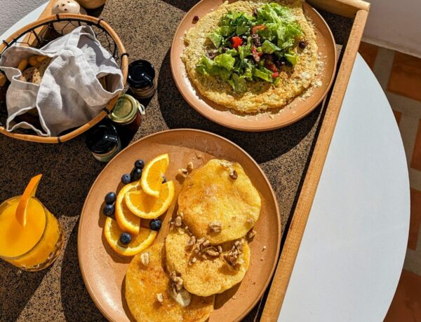 a round plate with mini vegan pancakes next to a vegan chickpea omelet on a tray with fresh juice and a bread basket in santorini