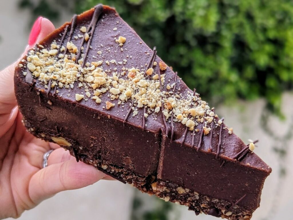 a single slice of dark chocolate hazelnut cheesecake held in a hand in front of greenery at a vegan bakery in athens greece