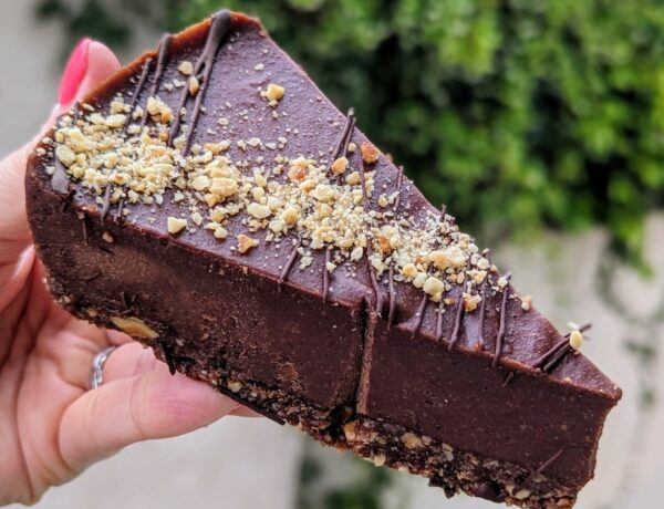a single slice of dark chocolate hazelnut cheesecake held in a hand in front of greenery at a vegan bakery in athens greece