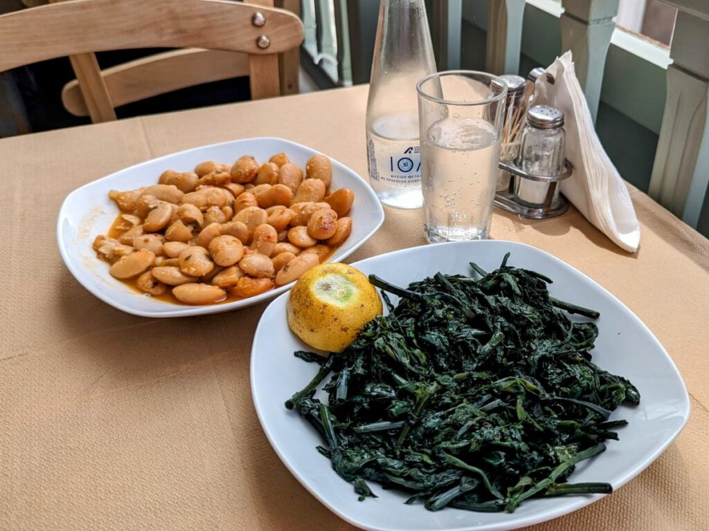 a white bowl filled with vegan giant beans in a tomatoes sauce next to a boiled green salad on a wooden table at Boulamatsis in naxos