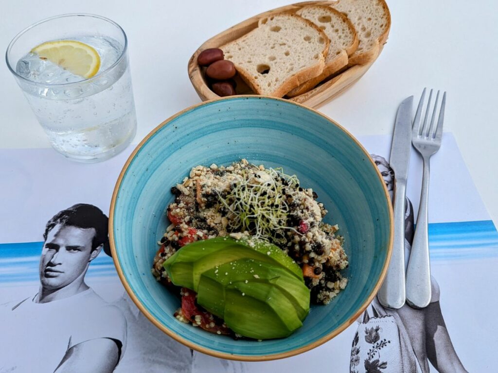 a light blue bowl on a white table with quinoa, veggies, and slices of avocado next to a basket of bread at nice n easy in mykonos