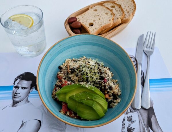a light blue bowl on a white table with quinoa, veggies, and slices of avocado next to a basket of bread at nice n easy in mykonos