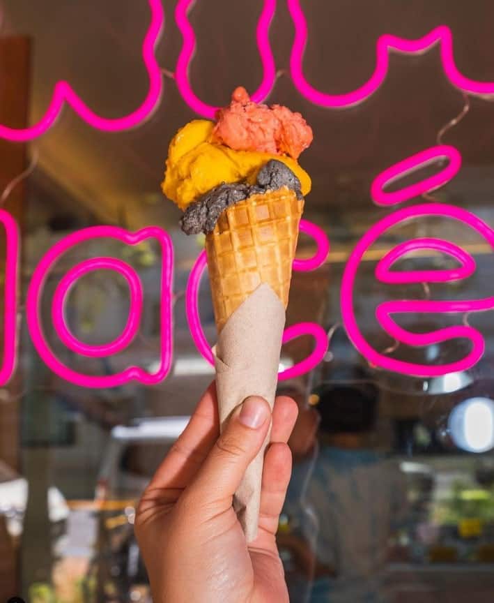 a single vegan ice cream cone with multiple scoops of strawberry and mango ice cream held in front of a neon sign in zurich