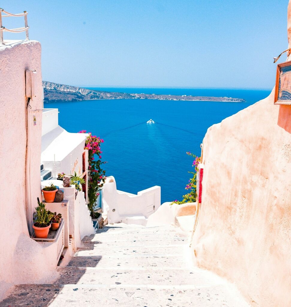 a white stone pathway between whitewashed building leading to the blue sea in greece