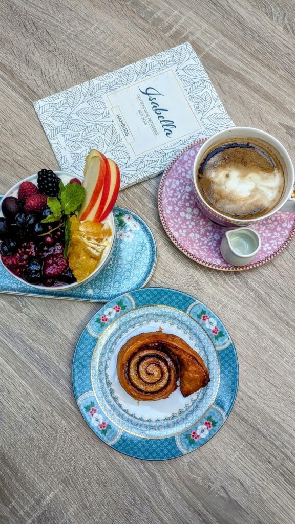 two small dishes filled with fruit and a golden cinnamon snail next to a coffee on a light wood table