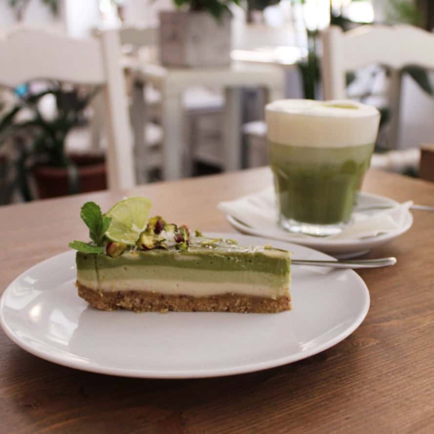 a slice of vegan matcha cheesecake with multiple layers of green filling next to a cup of matcha at my raw cafe in prague