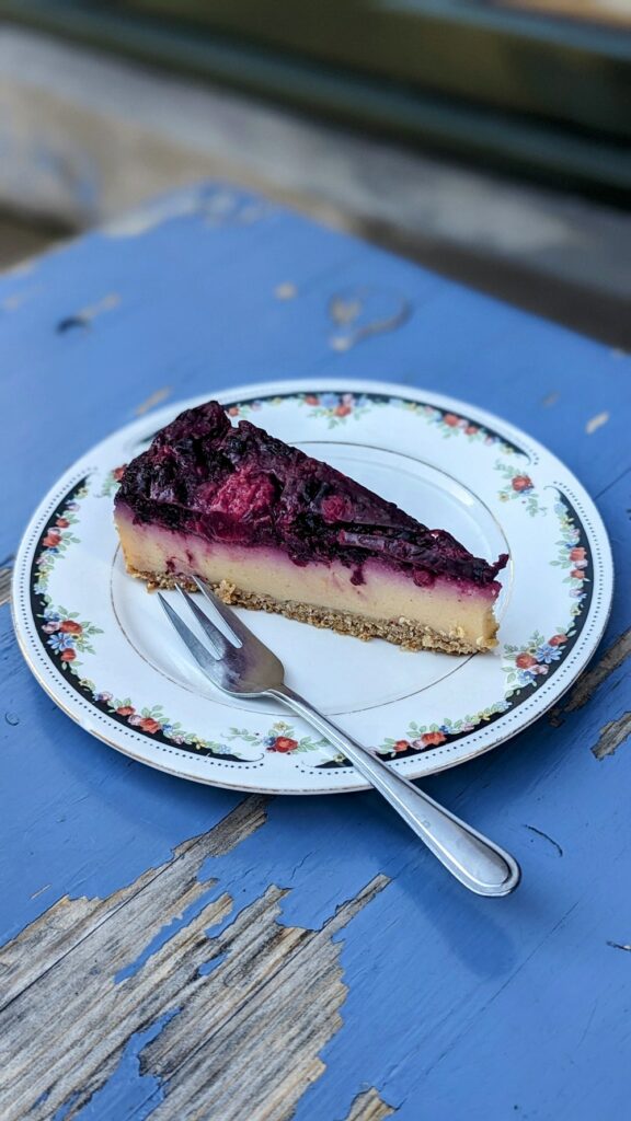 a small slice of vegan cheesecake topped with a dark berry topping on a white plate on a rustic blue table in hamburg