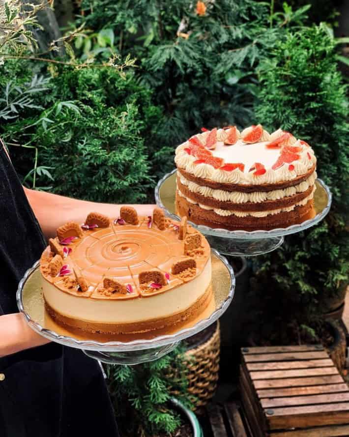 two large round vegan cakes topped with fresh fruit held up in the air in front of a green bush in prague