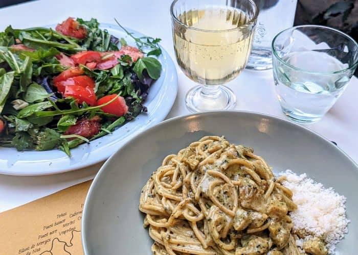 a bowl of creamy vegan pasta next to a bright green and red salad and a glass of white wine