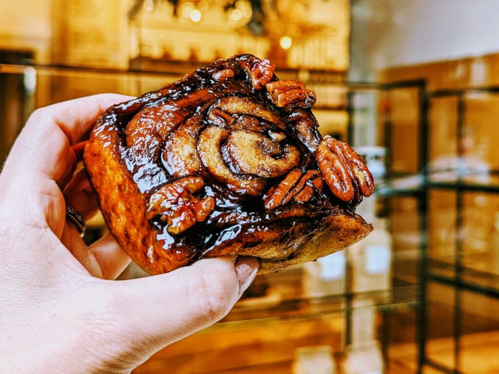 a person holding a vegan sticky bun in front of a bakery window in zurich