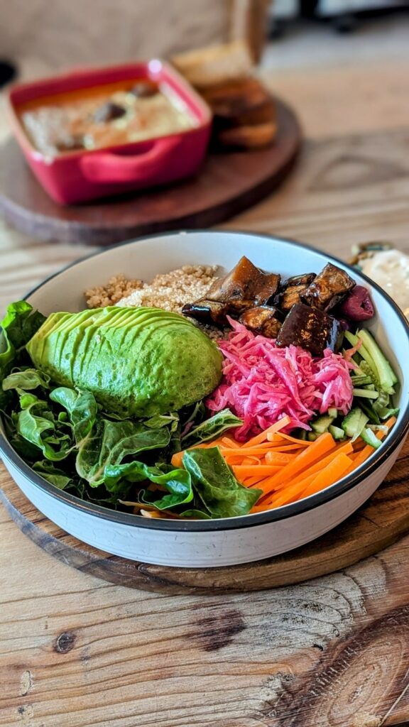 a large colorful bowl of fresh veggies on a wood table with a bowl of tomato soup in the background at the Conscious Kitchen in cape town 