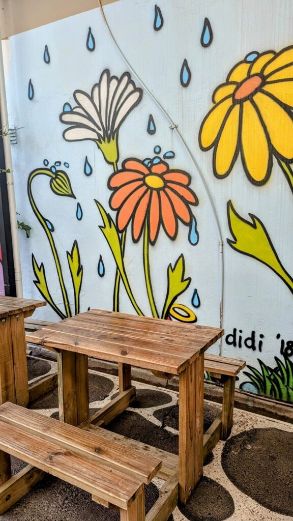 a colorful mural with giant orange, yellow, and white flowers behind mini picnic tables in an alleyway in cape town