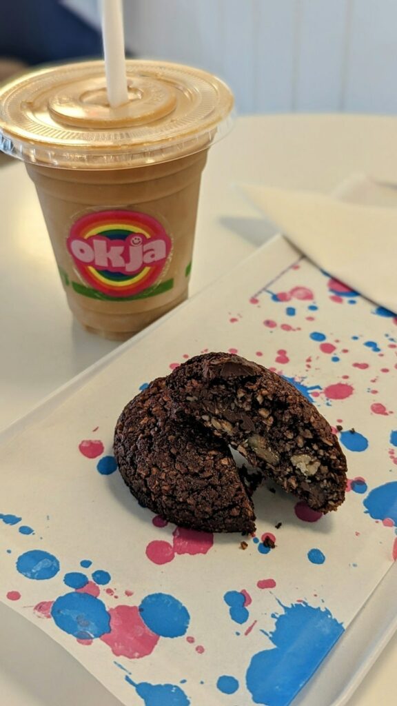 a dark chocolate and peanut butter vegan and gluten free cookie cut in half on a colorful piece of paper next to an oatmilk latte at okja in cape town