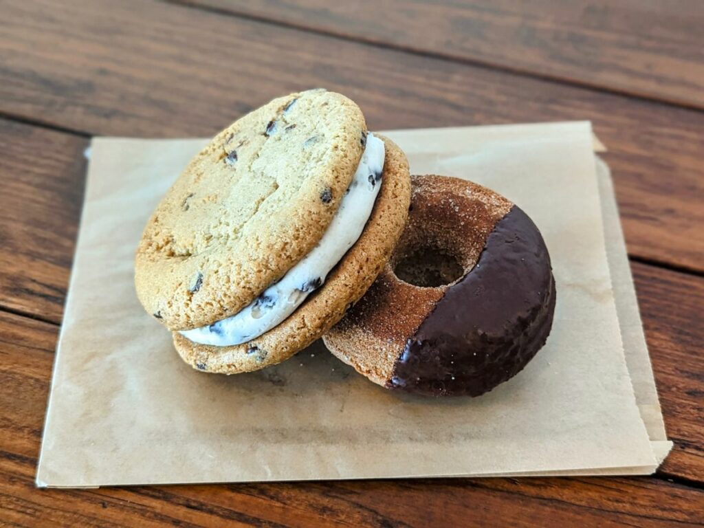 a vegan cookie sandwich leaning on a sugar donut thats been dipped partially in chocolate sitting on a square light brown bag