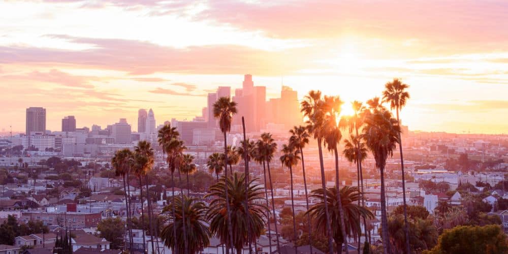 the LA skyline at sunrise with a pink and orange sky surrounding the buldings in downtown LA and in the foreground tall palm trees