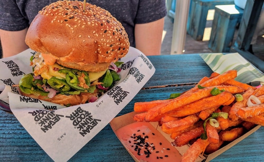 a giant vegan burger with cheese and veggies falling out next to a small tray of golden sweet potato fries with a small container of pink dipping sauce in amsterdam