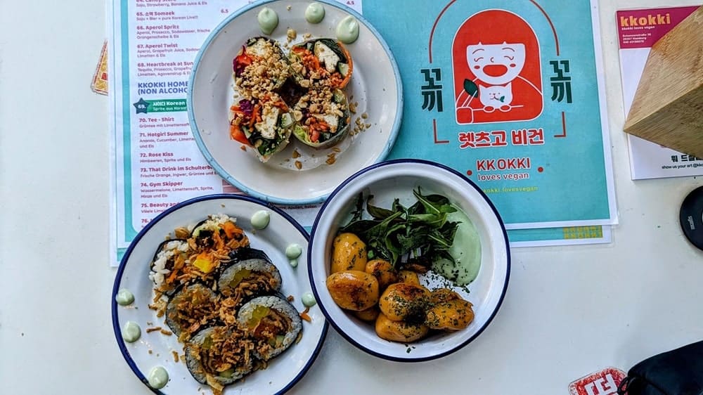 three small vegan korean dishes spread on a white table with a red and teal menu behind them in hamburg