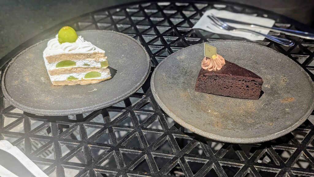 a slice of vegan spone cake with cream and giant green muscat grapes on a black plate next to a slice of dark chocolate cake on a black outdoor table at mercy vegan factory in osaka