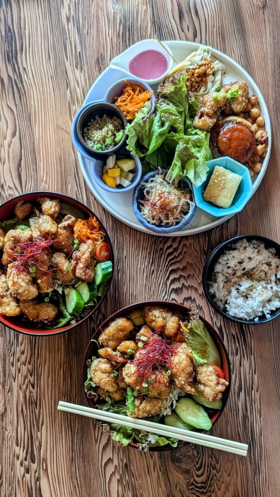 two bowls filled with golden vegan fried chick'n pieces with a little bit of greenery next to a larger round platter filled with little dishes of sauce and colorful veggies on a wood table at onwa in nara