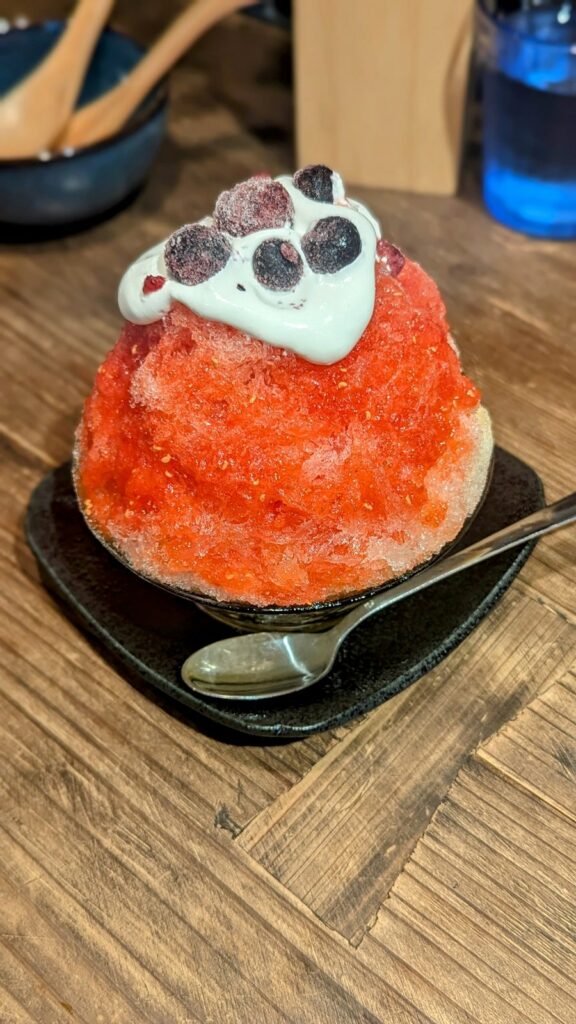 a giant vegan strawberry shaved ice covered in pink syrup and topped with vegan cream and a few strawberries on a wood table at vegan playpen in osaka