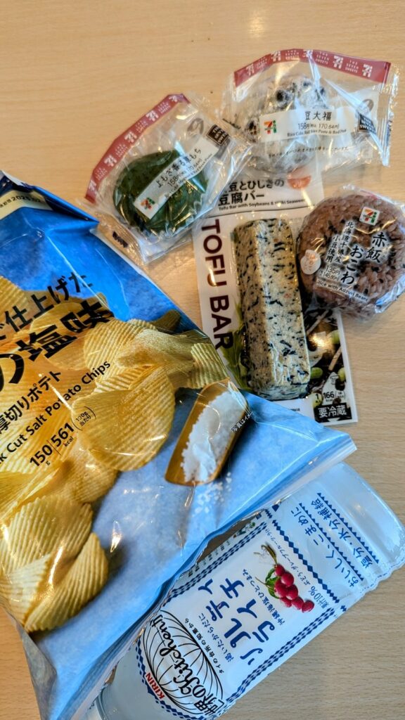 an array of vegan snack like a tofu bar, chips, two mochi, and a rice ball from a 7 eleven inside the haneda airport in japan