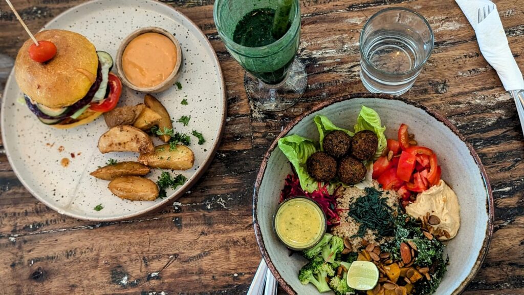 vegan burger on a plate with thick cut french fries next to a colorful veggie bowl with mini falafel on a wood table at the vegan restaurant alchemy in ubud, bali