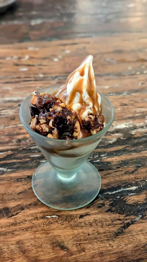 a small vegan ice cream sundae in a glass cup, topped with caramel sauce and chunks of dark chocolate sitting on a wood table at alchemy in ubud bali