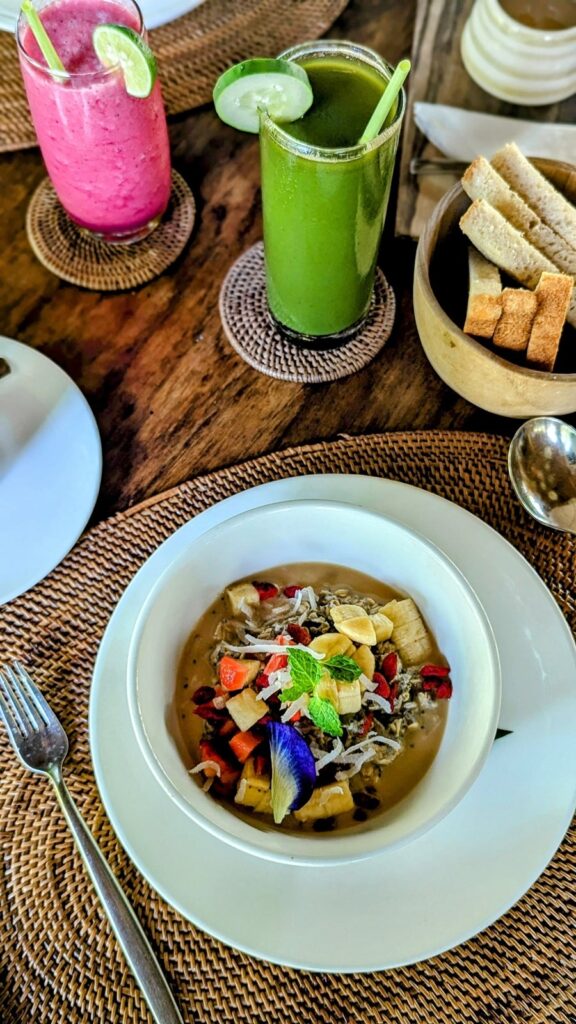 a small round white bowl of vegan porridge topped with dried and fresh fruit next to a green juice at fivelements retreat in ubud, bali