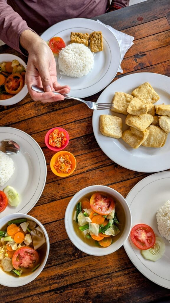 a spread of white dishes filled with golden fried tofu, tempeh, veggies, and small bowls of soup on a wood table with a single handle holding a fork in bali