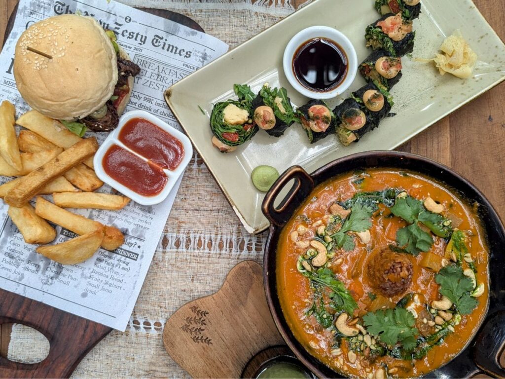 a spread of vegan lunch dishes at zest ubud - a large dark bowl filled with an orange peanut curry, next to mini colorful sushi rolls and a vegan burger with golden french fries on a newspaper style serving tray at zest ubud