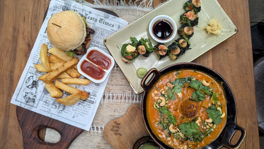 a spread of vegan lunch dishes at zest ubud - a large dark bowl filled with an orange peanut curry, next to mini colorful sushi rolls and a vegan burger with golden french fries on a newspaper style serving tray at zest ubud