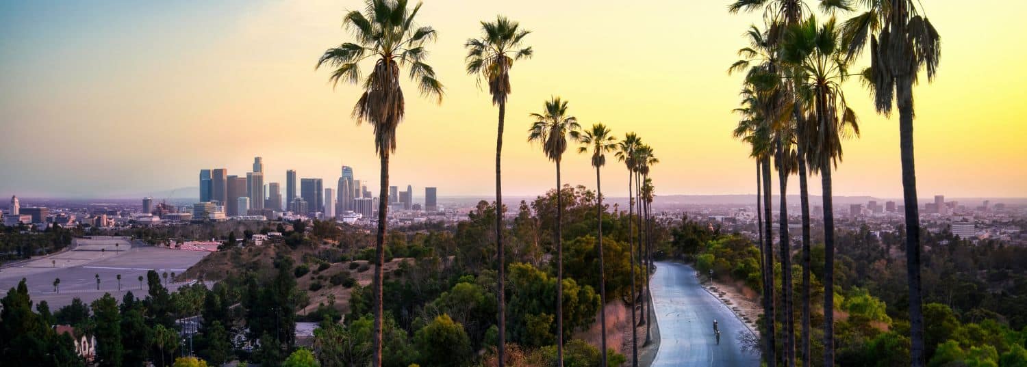 light pink and orange sky as the sun is setting over los angeles with palm trees surrounding an empty road