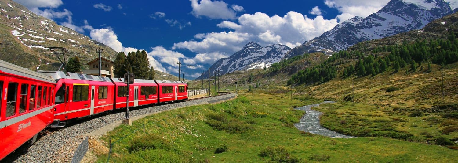 a red train car moving through the mountains of switzerland leaving behind green fields for snowcapped mountains