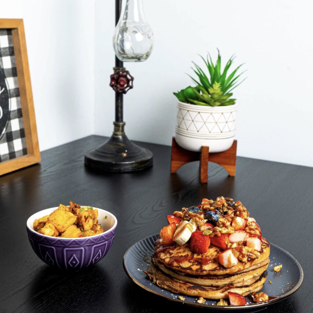 Plate of pancakes with a side of plantains. On a black table next to a succulent and a lamp
