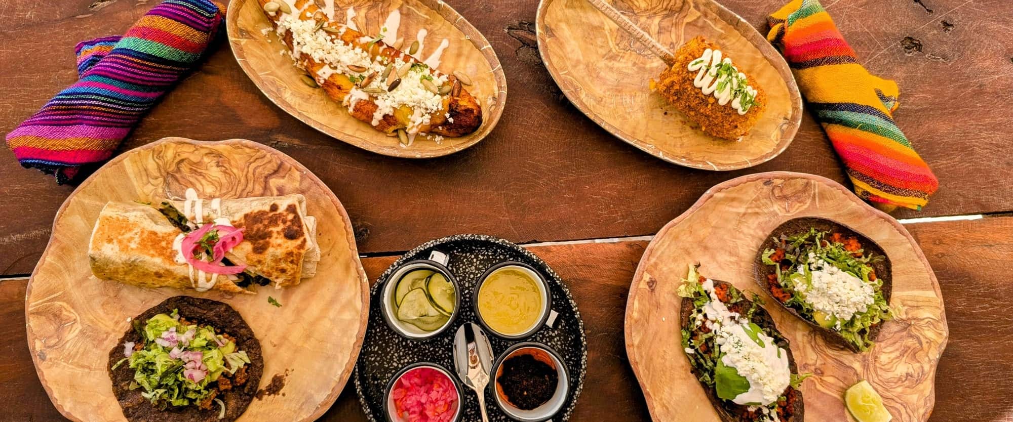 spread of vegan mexican food like tacos, corn, burritos, salsas, and more on a wood picnic table with colorful napkins