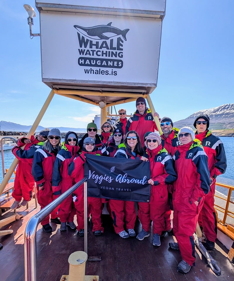 group photo of 15 people in bright red jump suits on a whale watching boat in iceland holding a sign that says veggies abroad vegan tours and travel