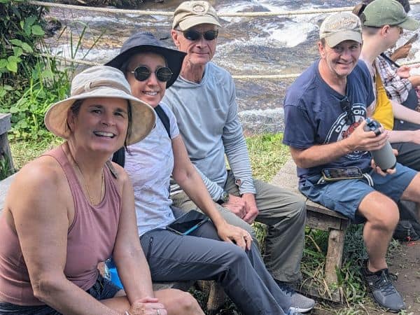 a group of smiling travelers sitting near a river waiting to see if elephants come down for a lunch snack in thailand
