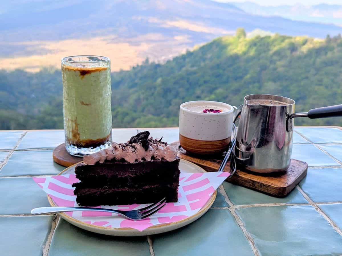 a single slice of dark chocolate vegan cake on a pink plate next to a green avocado iced coffee and a chai tea