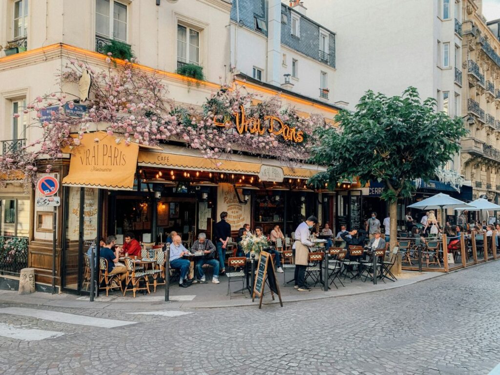 a small cafe on the corner of a street in paris with a yellow overhang and a large vine of flowers over the top