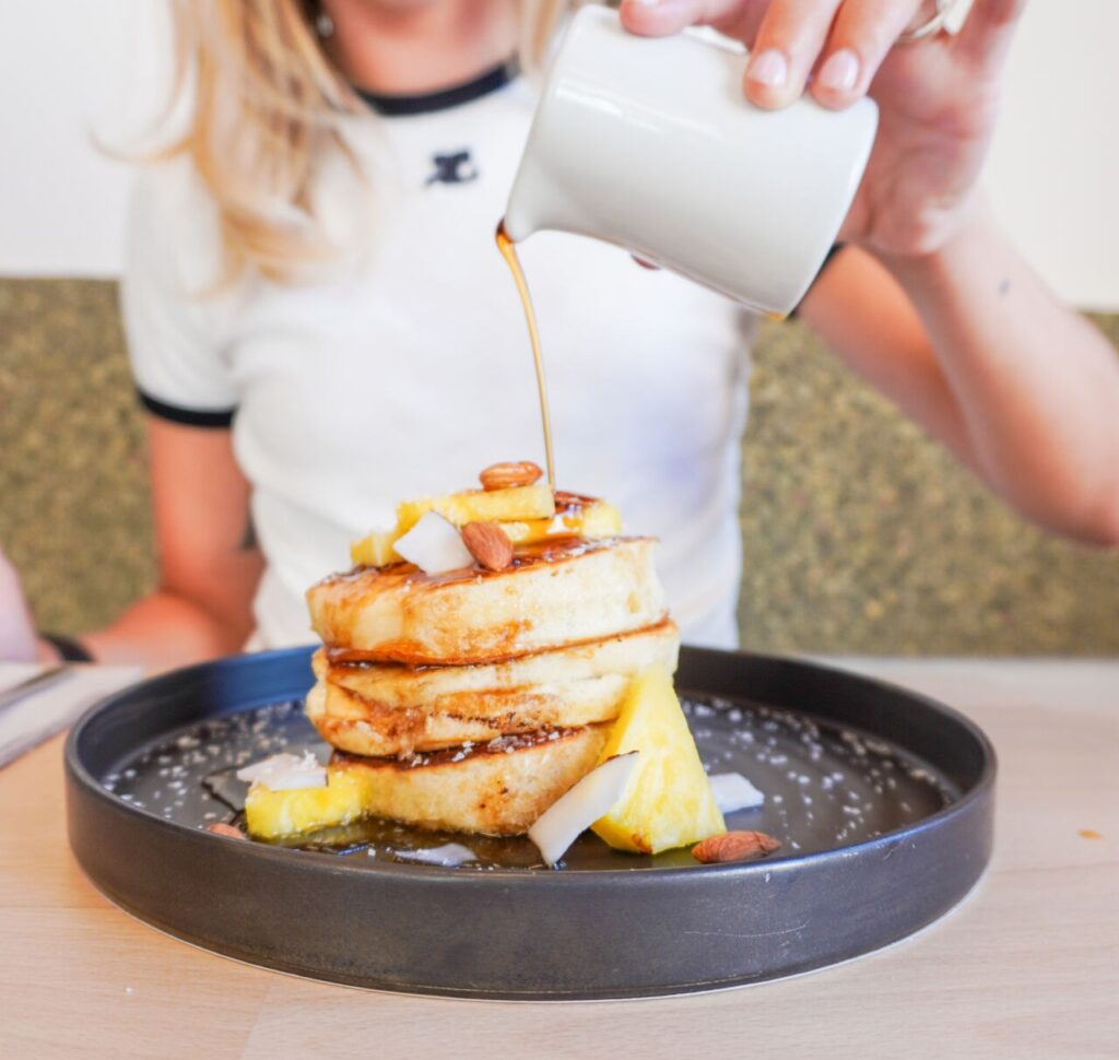 a stack of mini golden vegan pancakes being topped with maple syrup from a white syrup holder at bloomy rive gauche