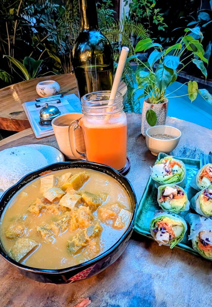 large bowl of vegan amok next to a bright orange juice and colorful rainbow rolls in Phnom Penh