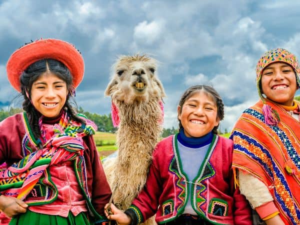 three Peruvian girls with a llama smiling and wearing brightly colored clothes
