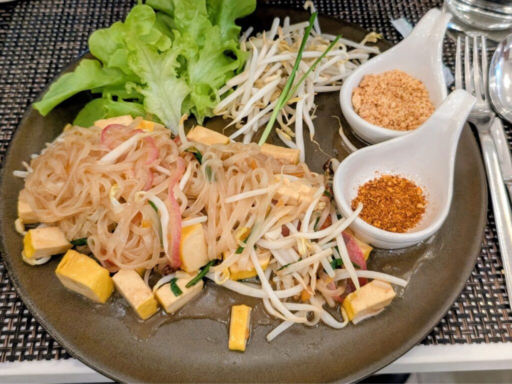 a black plate with vegan pad thai with tofu and little spoons with spices and peanuts in thailand