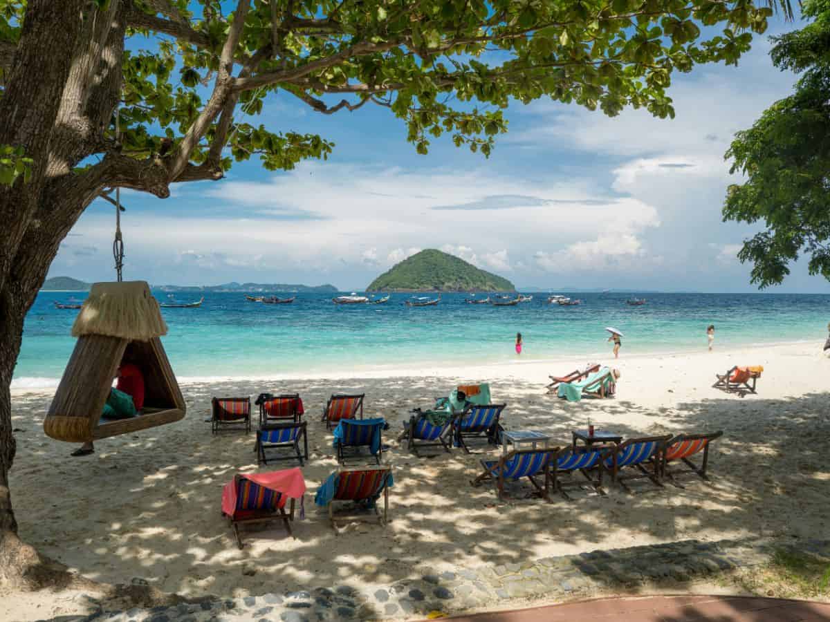 empty beach chairs under a shady tree on a white sand beach looking out onto clear blue water in thailand