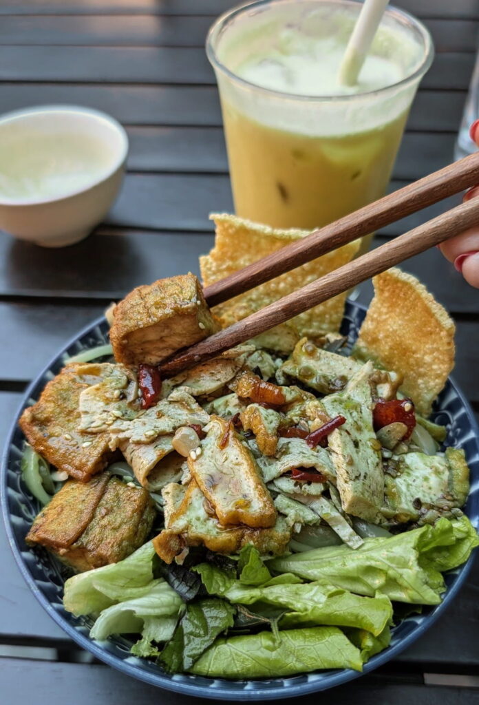 chopsticks grabbing a piece of tofu out of a noodle bowl with green leafy lettuce and crispy chips in hoi an